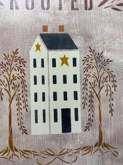 close up of a rustic painted sign or wall art featuring a white house with a dark roof, two gold stars, and brown trees on a textured white-gray-brown background.