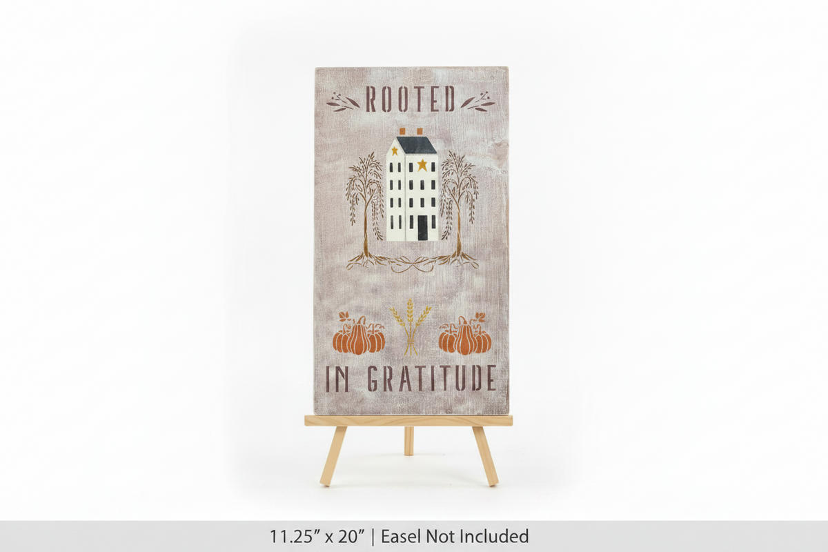 A vertical rectangular decorative sign on a small wooden easel stand, featuring the words 'ROOTED IN GRATITUDE' with an illustration of a house, trees, pumpkins, and wheat on a distressed beige background.