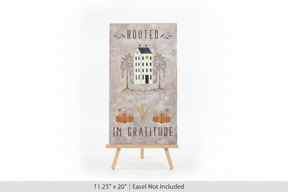 A vertical rectangular decorative sign on a small wooden easel stand, featuring the words 'ROOTED IN GRATITUDE' with an illustration of a house, trees, pumpkins, and wheat on a distressed beige background.