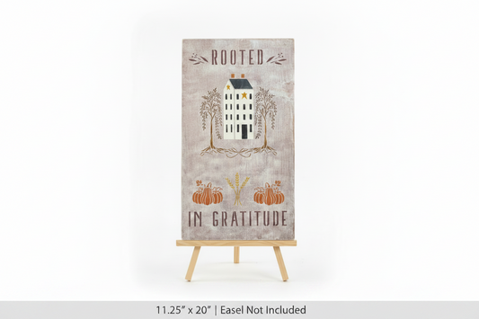 A vertical rectangular decorative sign on a small wooden easel stand, featuring the words 'ROOTED IN GRATITUDE' with an illustration of a house, trees, pumpkins, and wheat on a distressed beige background.