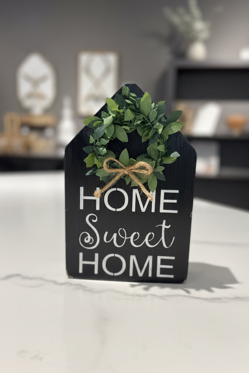 A black wooden wall sign with the words 'HOME sweet HOME' in white script and block lettering, decorated with a green leafy wreath and tied with a jute twine bow, shown against a white wall.