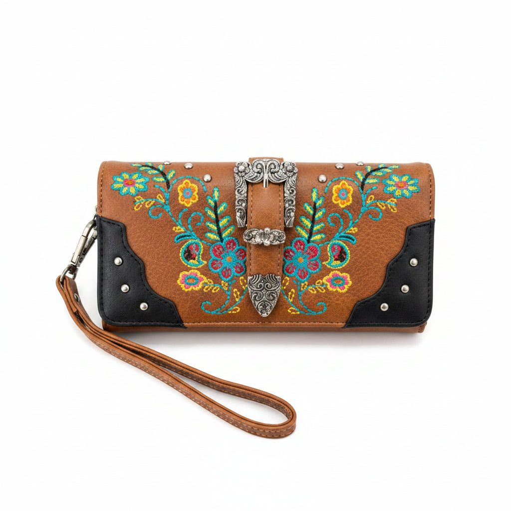 A western-style wristlet clutch wallet with a tan leather body, black leather corner panels, and colorful floral embroidery. It features a silver-tone concho buckle closure, silver studs, and a matching tan wrist strap