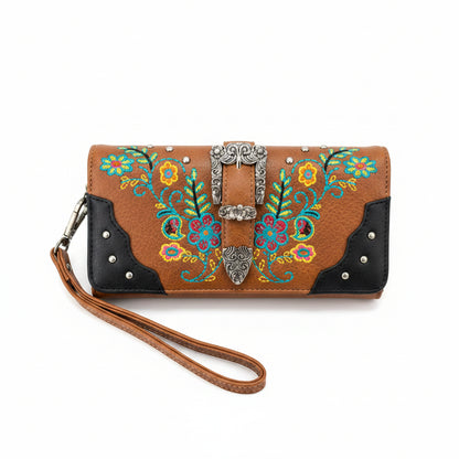 A western-style wristlet clutch wallet with a tan leather body, black leather corner panels, and colorful floral embroidery. It features a silver-tone concho buckle closure, silver studs, and a matching tan wrist strap