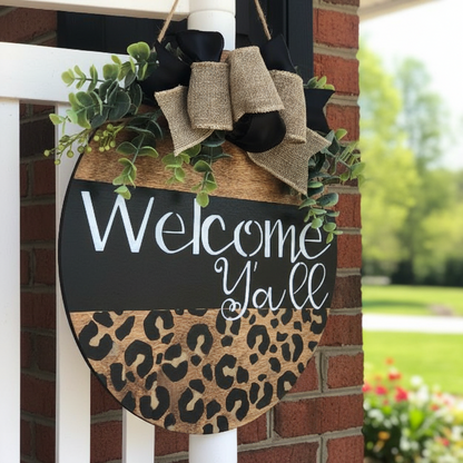 A 12-inch round door hanger with a cheetah print design, featuring the text 'Welcome Y'all' in bold script, accompanied by a handmade bow with faux greenery.