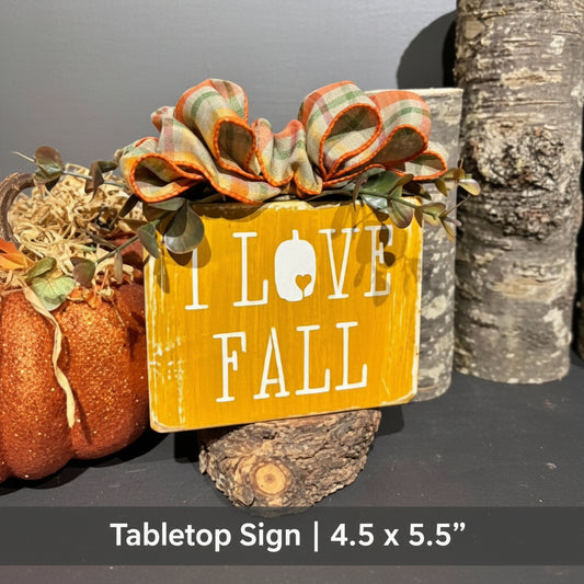 A decorative wooden sign with the words 'I LOVE FALL' in white lettering on a yellow-orange background, styled with faux autumn leaves, pinecones, and small wood pieces against a dark gray backdrop.