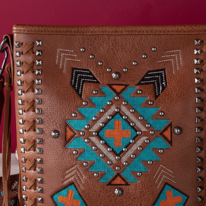 A close-up of a brown leather shoulder bag featuring a Southwestern geometric pattern in turquoise, orange, black, and white, embellished with silver studs and a braided strap detail on the side.