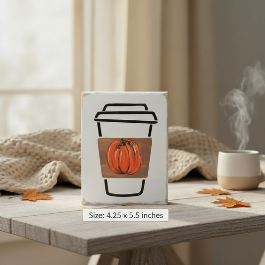 A small decorative sign with a painted coffee cup design featuring an orange pumpkin, styled on a wooden table with a knitted blanket, scattered autumn leaves, and a steaming mug in the background.